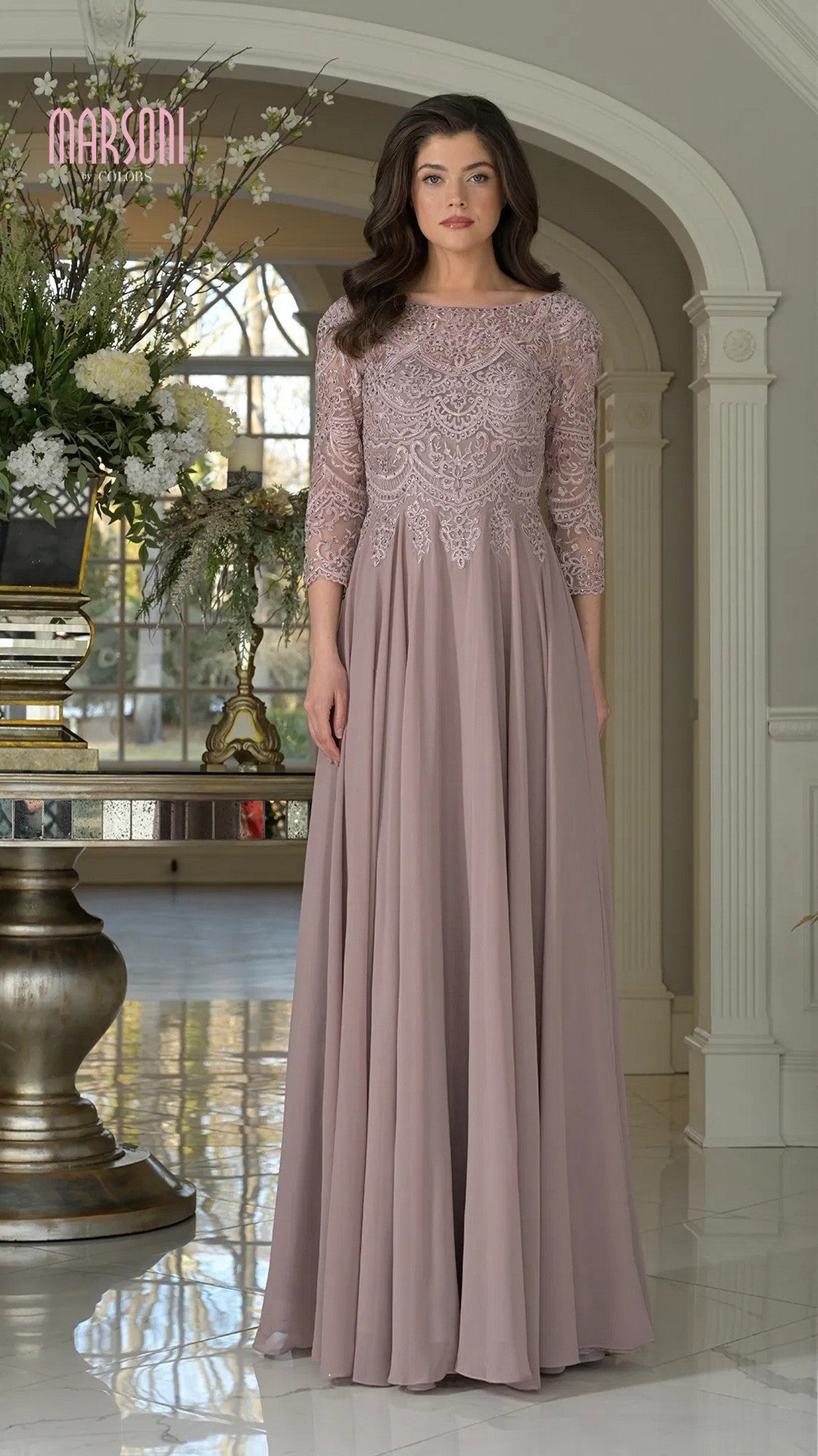 A woman stands indoors wearing the Marsoni M238SL, a long formal dress with lace bodice detailing, three-quarter sleeves, and a flowing chiffon A-line skirt in mauve.